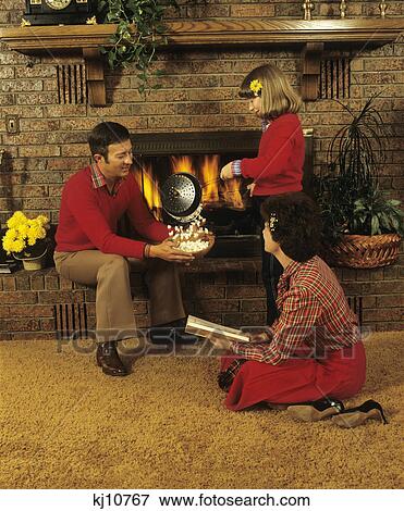 Stock Photo - 1970S Family Making Popcorn Fireplace Mother Reading Book Father Daughter Cooking. Fotosearch