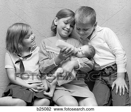 Stock Image - 1970S Girl Feeding Bottle To Baby Sister With Brother & Sister Sitting On Either Side Of Her Looking On. Fotosearch
