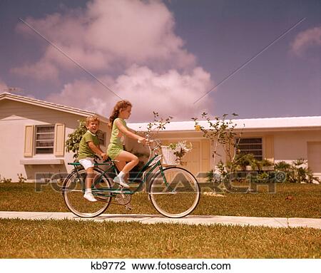 Stock Image - 1970S Girl Riding Bike On Suburban Street Boy Rides Piggy Back . Fotosearch