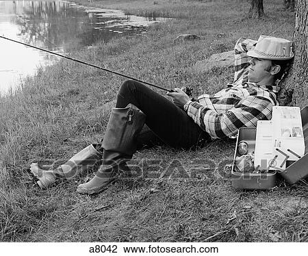 Stock Image - 1970S Man Sleeping Against A Tree Trunk While Fishing In A Lake. Fotosearch
