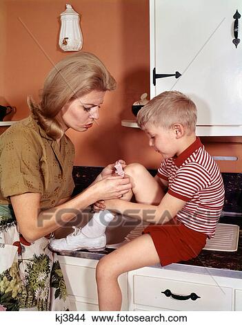 Picture - 1970S Mother Giving First-Aid Putting Band-Aid On Knee Of Son In Kitchen . Fotosearch