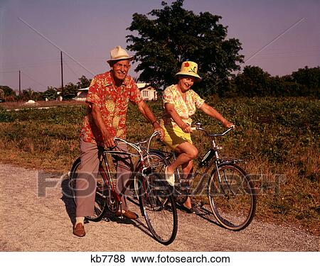 Stock Photo - 1970S Senior Elderly Retired Couple Riding Bikes Wearing Straw Hats Hawaiian Print Shirts . Fotosearch