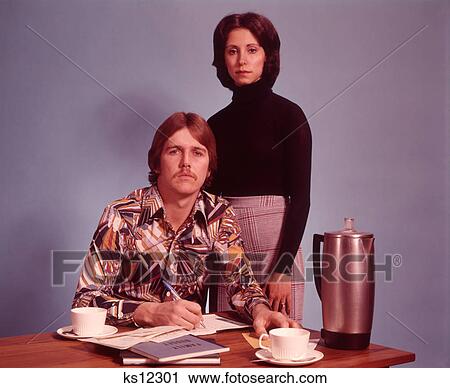 1970S Serious Couple Husband Sitting Wearing Loud Print Shirt Writing Family Budget Wife Standing Behind Coffee Pot On Desk View Large Photo Image Stock Image - 1970S Serious Couple Husband Sitting Wearing Loud Print Shirt Writing Family Budget Wife Standing Behind Coffee Pot On Desk. Fotosearch