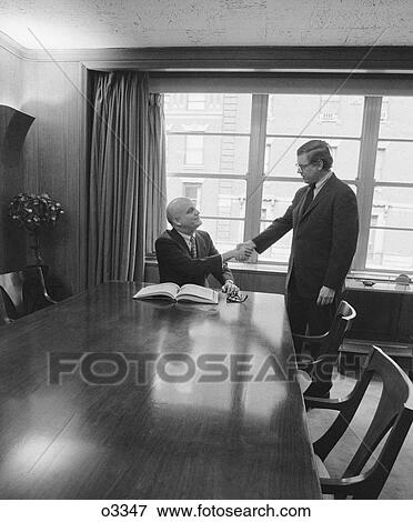 Stock Photo - 1970S Two Men One Seated Shaking Hands Long Wooden Conference Table. Fotosearch