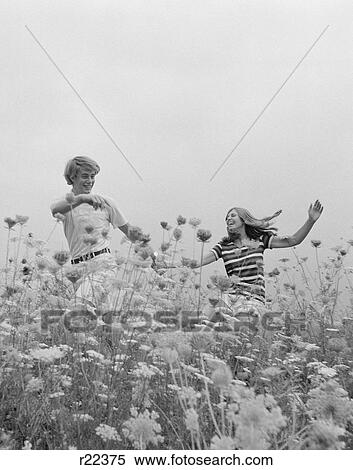 Stock Photography - 1970S Young Couple Holding Hands Running Through Field Of Flowers. Fotosearch