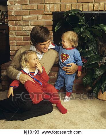 Stock Image - 1980S Father Sitting By Fireplace With Two Young Children In Pajamas. Fotosearch