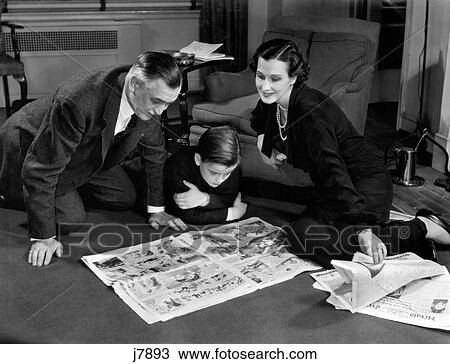 Stock Image - 1930S 1940S Father And Mother Reading Sunday Newspaper Comics With Son Sitting On Living Room Floor Together. Fotosearch