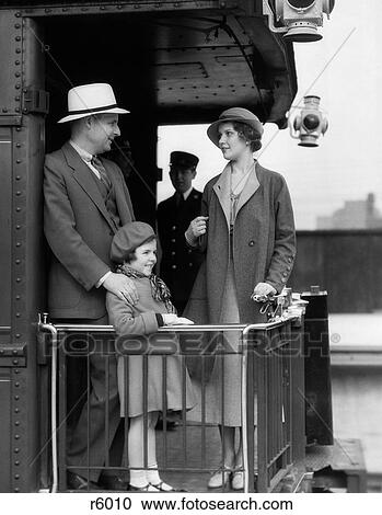 Stock Image - 1930S Family Of Three Standing At Railing At Back Of Train Car With Porters In Background. Fotosearch