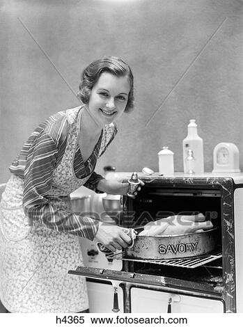 Stock Photography - 1930S Housewife In Apron Taking Turkey In Pan Reading Savory Out Of Oven Looking At Camera. Fotosearch