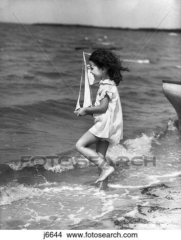 1930S Side View Of Little Girl In White Dress Walking Out Into Surf Holdingtoy Sailboat View Large Photo Image Picture - 1930S Side View Of Little Girl In White Dress Walking Out Into Surf Holdingtoy Sailboat. Fotosearch