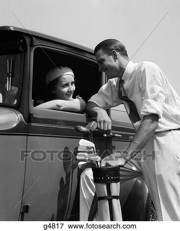 Stock Photo - 1930S Woman In Driver'S Seat Of Car Leaning Out Of Window Talking To Man Standing Holding Golf Bag. Fotosearch