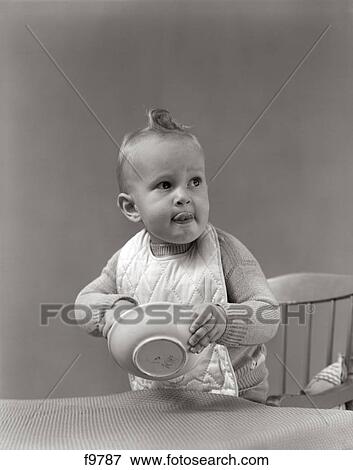 Stock Photo - 1940S Baby Wearing Bib Licking Lips & Putting Hand In Cereal Bowl. Fotosearch