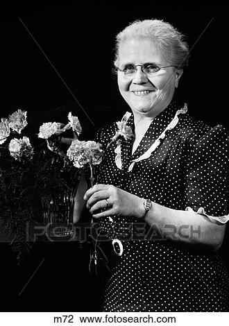 Stock Image - 1940S Elderly Woman Putting Carnation Flowers In Vase Looking At Camera. Fotosearch