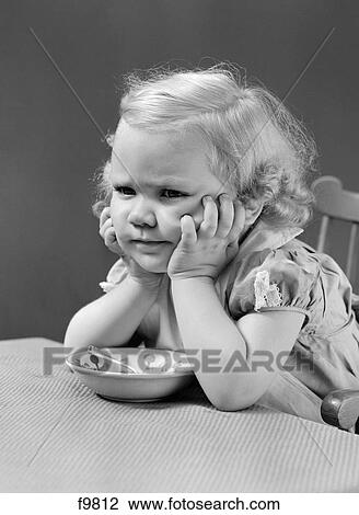 Stock Image - 1940S Girl At Table With Head Cradled In Both Hands Frown On Her Face A Cereal Bowl And Spoon Between Her Elbows On Table. Fotosearch