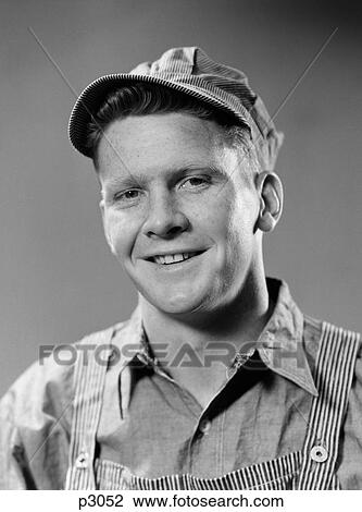 Stock Image - 1940S Portrait Of Smiling Blue Collar Working Man Wearing Overalls And Engineer Hat Looking At Camera. Fotosearch