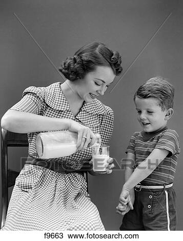 Stock Image - 1940S Woman Mother Sitting Pouring Glass Of Milk For Boy Son Standing Beside Her. Fotosearch