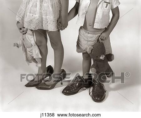 Stock Photo - 1950S 1960S Close-Up Of Little Girl & Boy From Neck Down Wearing Parents' Shoes. Fotosearch