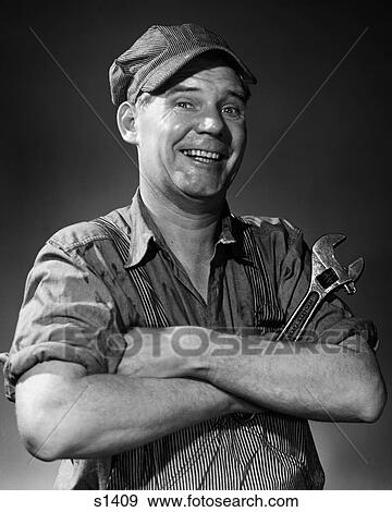Stock Photo - 1950S 1960S Smiling Portrait Man In Striped Cap & Overalls Crossing Arms Holding Monkey Wrench Looking At Camera. Fotosearch