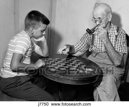 Stock Image - 1950S Boy Playing Checkers With Grandfather Smoking Pipe & Picking Up Piece To Jump His Checker. Fotosearch