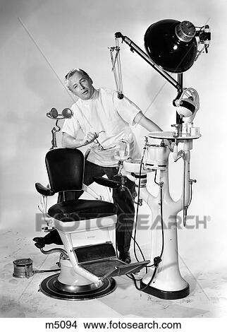1950S Man Dentist Holding Glasses Speaking Foot Propped Up On Dental Chair And Equipment Looking At Camera View Large Photo Image Picture - 1950S Man Dentist Holding Glasses Speaking Foot Propped Up On Dental Chair And Equipment Looking At Camera. Fotosearch