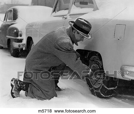Stock Photo - 1950S Man In Coat & Hat Kneeling Down In Snow To Adjust Chains On Tires. Fotosearch