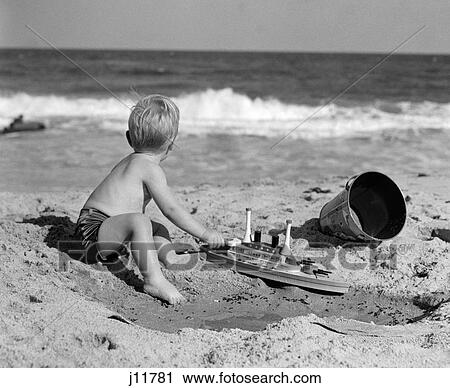 Stock Image - 1950S Side View Of Boy Playing In Sand With Bucket & Toy Boat Head Turned Toward Water. Fotosearch