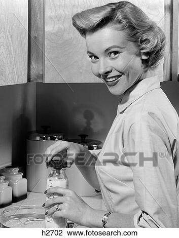 Stock Image - 1950S Smiling Blonds Woman Mother Warming A Baby Bottle Of Milk In Kitchen Looking At Camera. Fotosearch