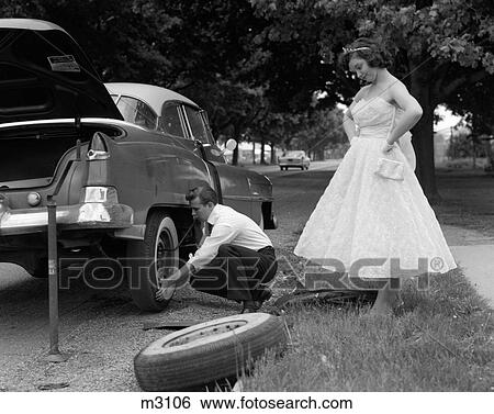 1950S Teenage Boy Crouching Down Changing Flat Tire Prom Date Watching With Hands On Hips View Large Photo Image Stock Photograph - 1950S Teenage Boy Crouching Down Changing Flat Tire Prom Date Watching With Hands On Hips. Fotosearch