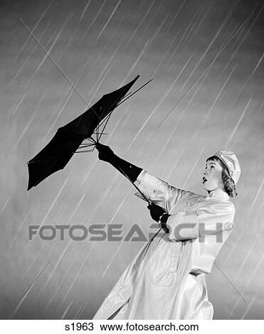 Stock Image - 1950S Woman In Rain Coat Trying To Use Broken Umbrella In The Wind. Fotosearch