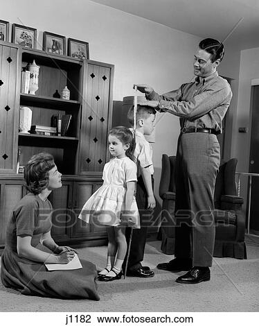 Stock Image - 1960S Father Measuring Daughter & Son Back-To-Back With Mother Recording Information. Fotosearch