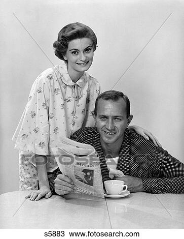 Stock Image - 1960S Husband Wife Wearing Pajamas At Breakfast Table Woman Behind Man Holding Newspaper Both Smiling And Looking At Camera. Fotosearch