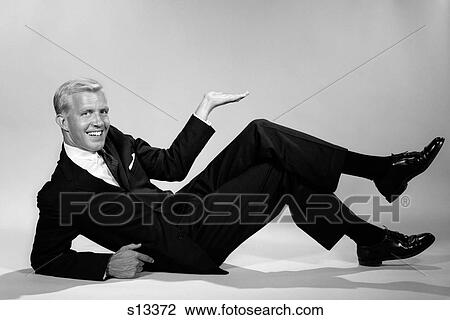 Stock Image - 1960S Smiling Businessman Reclining Propped On Elbow Holding Hand Up In Air Looking At Camera. Fotosearch