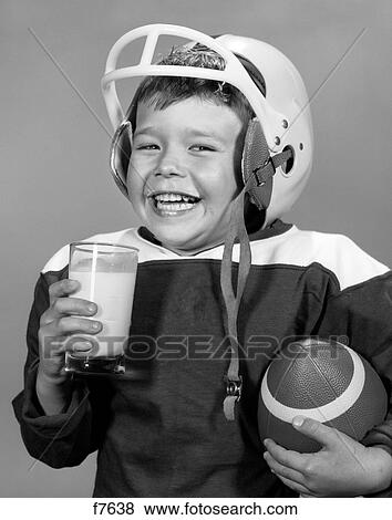 Stock Photo - 1960S Smiling Young Boy With Milk Mustache In Football Uniform & Helmet Ball Under Arm Holding Glass Of Milk Looking At Camera. Fotosearch