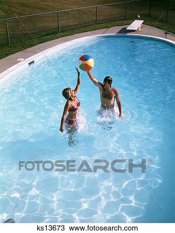Stock Image - 1980 1980S Couple Man Woman Playing Jumping Up After Beach Ball Backyard In Ground Swimming Pool . Fotosearch