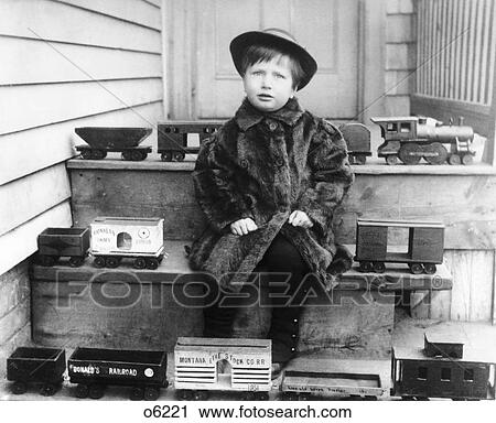Stock Image - 1880S 1890S 1900S Boy Wearing Fur Coat Sitting On Steps Surrounded By Toy Railroad Trains Looking At Camera. Fotosearch