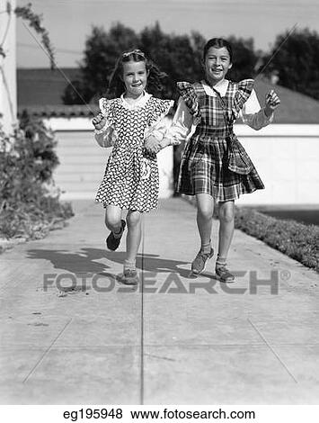 Stock Photo - 1930S 1940S Two Girls Skipping Down Sidewalk Holding Hands Looking At Camera. Fotosearch