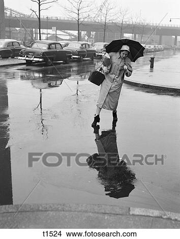 1950S Woman In Rain Coat Hat Boots Holding Umbrella Handbag Stepping To Avoid Puddles While Crossing City Street In Foul Weather View Large Photo Image Picture - 1950S Woman In Rain Coat Hat Boots Holding Umbrella Handbag Stepping To Avoid Puddles While Crossing City Street In Foul Weather. Fotosearch
