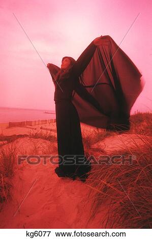 1960S 1970S Woman In Eerie Red Light Standing On Sand Dune Cape Blowing In Wind Like Wings View Large Photo Image Stock Photo - 1960S 1970S Woman In Eerie Red Light Standing On Sand Dune Cape Blowing In Wind Like Wings. Fotosearch
