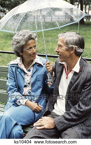 1970S Elderly Couple Sitting On Park Bench Holding An Umbrella View Large Photo Image Picture - 1970S Elderly Couple Sitting On Park Bench Holding An Umbrella . Fotosearch