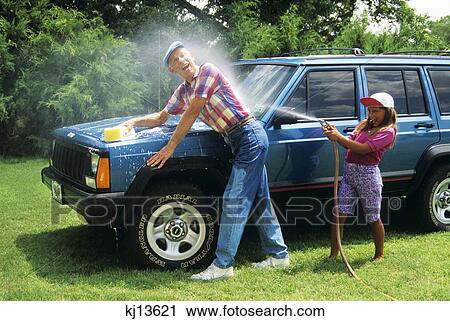Stock Image - 1980S Grandfather Washing Car Granddaughter Spraying Him With Water From Hose. Fotosearch