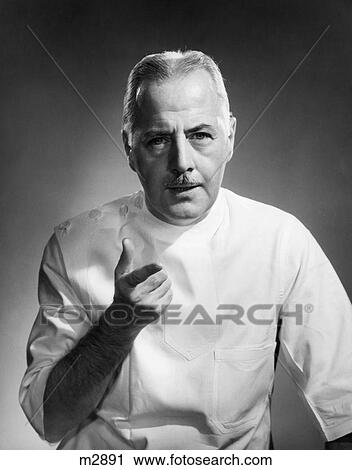 Stock Image - 1940S Mature Man Doctor White Tunic Pointing Looking At Camera. Fotosearch