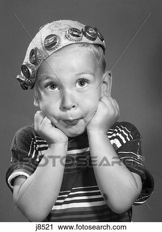 Stock Image - 1950S Portrait Of Boy Resting Head On Fists Looking At Camera Wearing Beanie Hat Decorated With Soda Bottle Caps. Fotosearch