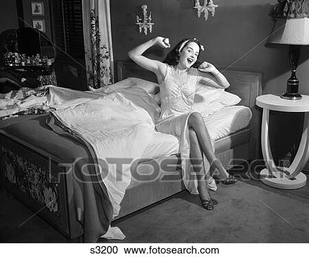Stock Image - 1950S Smiling Young Woman In Nightgown Slippers Sitting Edge Bed Looking At Camera Yawning Stretching Waking Up. Fotosearch