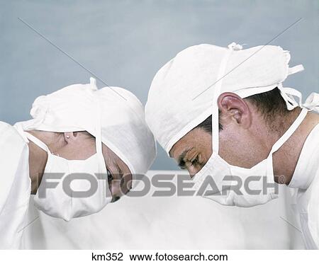 Stock Image - 1960S Couple Man Woman Doctor & Nurse White Surgical Cap & Mask Head To Head In Profile Operationg Looking Down . Fotosearch