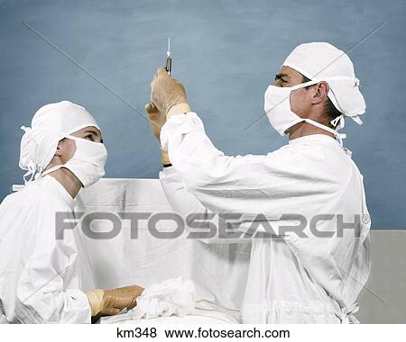 Stock Photo - 1960S Man Doctor Woman Nurse Wearing White Masks Caps Gowns Sterile Rubber Gloves Preparing Injection Syringe. Fotosearch