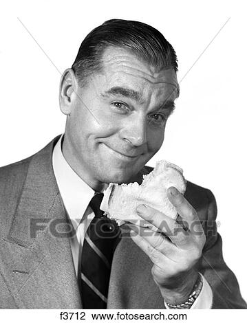 Stock Image - 1960S Smiling Businessman Wearing Suit And Tie Eating Sandwich Looking At Camera. Fotosearch