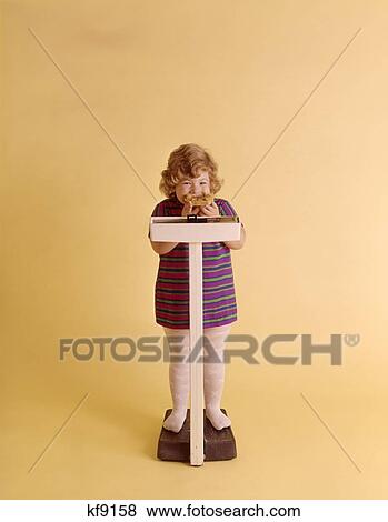 Stock Photo - 1970S Chubby Blonde Girl Striped Dress Eating Peanut Butter On Bread Standing On Scale. Fotosearch