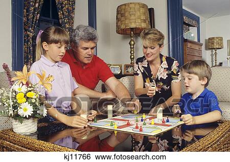 Stock Photograph - 1980S 1990S Engaged Family Of Four Playing Parcheesi Board Game At Home Sitting In Living Room. Fotosearch