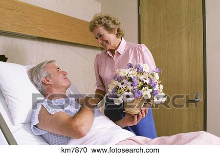Stock Image - 1980S Woman Giving Flowers To Hospitalized Man Lying In Bed. Fotosearch