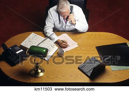 Stock Image - 1990S Overhead View Of Senior Doctor Sitting At Desk Talking On Telephone And Writing On Chart . Fotosearch